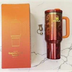 May include: A Stanley Quencher 112.0 Tumbler in a gradient red and orange color scheme, with a matching handle and lid. The tumbler has a floral design and the Stanley logo. The box next to it has a matching design and the text "Quencher 112.0 Tumbler 40 oz (1.1L)".