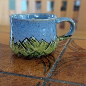 May include: A handmade ceramic mug with a blue sky and mountain design. The mug features a light blue glaze on the upper half, transitioning to green at the base. Black lines depict the mountain range. The handle is also blue and green.