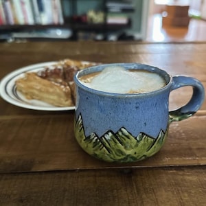 Pode incluir: Uma caneca de cerâmica com uma seção superior azul e um design de cordilheira verde na base, cheia de uma bebida de café coberta com espuma branca. A caneca apresenta uma alça curva e um acabamento de esmalte salpicado na superfície.