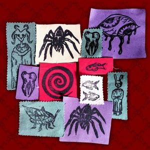 Puede incluir: Una colección de parches de tela rectangulares con diseños serigrafiados en tinta negra que incluyen arañas, dientes, una espiral, peces, un saltamontes y figuras con cuernos u orejas de conejo sobre fondos azul claro, morado, rojo y blanco.
