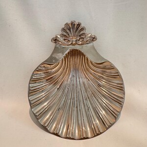 May include: A silver-toned, shell-shaped decorative dish. The dish has a detailed, ridged design with a decorative flourish at the top. The interior has a gold-toned finish, and the dish is set against a neutral background.