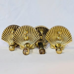 May include: A set of four gold-coloured metal wall hooks shaped like scallop shells. Each hook features a fan-like ribbed design with two small decorative spheres near the top and a larger spherical knob at the base for hanging items.