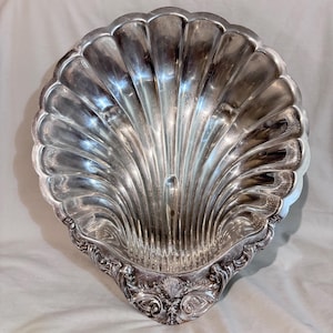 May include: A large, polished silver-toned serving dish shaped like a scallop shell. The interior features deep, radiating fluted grooves, while the base is decorated with intricate, ornate scrollwork and a small, central shell motif detail.