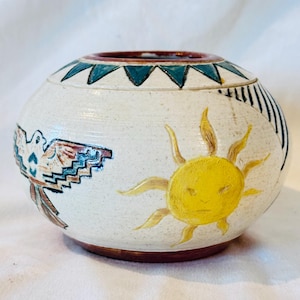 May include: A round ceramic bowl with a speckled off-white glaze. The rim features a teal triangle pattern. The body displays a hand-painted yellow sun, a stylized bird with brown and blue geometric wings, and dark blue diagonal line accents.