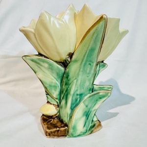 Könnte beinhalten: Eine Keramikvase in Form eines Bündels blassgelber Tulpenblüten mit markanten grünen Blättern und Stängeln. Der Sockel der Vase ist braun und strukturiert und stützt die florale Struktur, die eine glänzende Oberfläche und geformte Details aufweist.