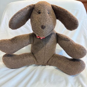 May include: A brown plush dog toy with a textured, fuzzy surface, long floppy ears, and dark bead eyes. The stuffed animal features a small, dark brown stitched nose and a decorative red and white floral patterned fabric collar around its neck.