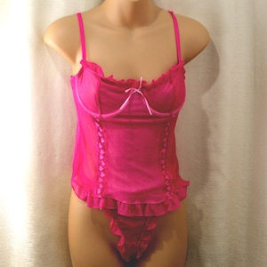 May include: A bright pink camisole and thong set featuring sheer mesh fabric, thin shoulder straps, and ruffled trim along the neckline and hem. The bodice includes vertical ribbon detailing with small heart accents and a center bow.