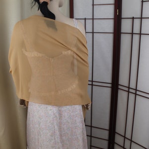 May include: A gold sheer shawl draped over a white dress with a floral pattern. The shawl is made of a lightweight fabric and has a delicate drape.
