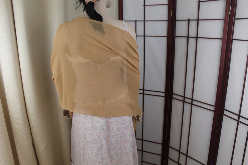 May include: A gold sheer shawl draped over a white dress with a floral pattern. The shawl is made of a lightweight fabric and has a delicate drape.
