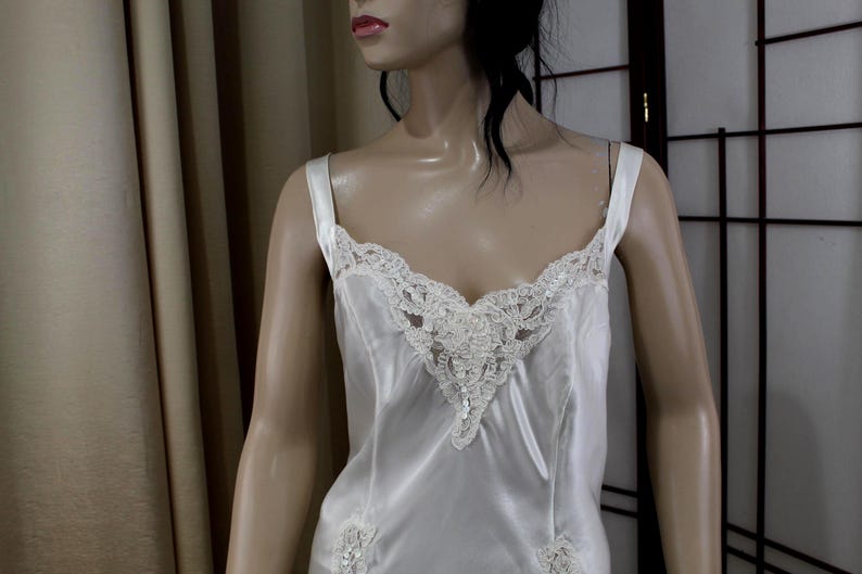 Puede incluir: Camis&oacute;n de sat&eacute;n color marfil con delicados adornos de encaje. El camis&oacute;n presenta un escote en V y tirantes finos. Los detalles de encaje se concentran alrededor del escote y en el dobladillo. La prenda se exhibe en un maniqu&iacute;.