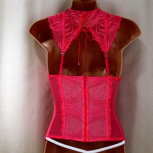 K&ouml;nnte beinhalten: Ein pinkfarbenes Korsett mit einem hohen, mit Spitze besetzten Kragen und verstellbaren Tr&auml;gern. Die R&uuml;ckseite hat einen Rei&szlig;verschluss und ein zartes Spitzendesign. Unten ist eine wei&szlig;e Verzierung zu sehen.