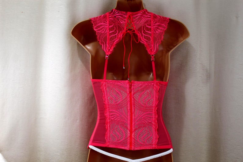 K&ouml;nnte beinhalten: Ein pinkfarbenes Korsett mit einem hohen, mit Spitze besetzten Kragen und verstellbaren Tr&auml;gern. Die R&uuml;ckseite hat einen Rei&szlig;verschluss und ein zartes Spitzendesign. Unten ist eine wei&szlig;e Verzierung zu sehen.