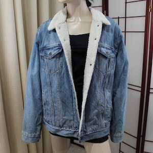 Könnte beinhalten: Hellblaue Jeansjacke mit weißem Sherpa-Futter und -Kragen. Die Jacke hat zwei Brusttaschen und Knopfverschlüsse. Ein klassisches, lässiges Oberbekleidungsstück.