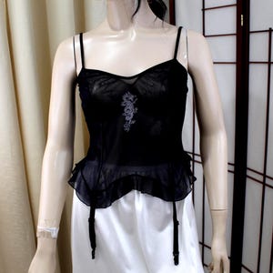 May include: A black sheer camisole featuring thin shoulder straps, a sweetheart neckline, and a delicate silver dragon embroidery centered on the chest. The garment includes a ruffled peplum hem and four attached black garter straps hanging down.