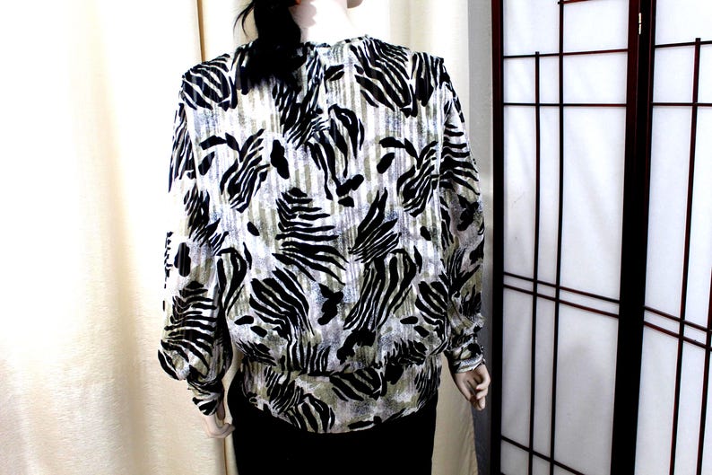 Pu&ograve; includere: Una camicetta a maniche lunghe con un motivo animalier bianco e nero simile a quello di una zebra su uno sfondo testurizzato verde oliva e grigio. Il capo presenta un orlo e polsini elasticizzati arricciati, con scollo rotondo e vestibilit&agrave; ampia.
