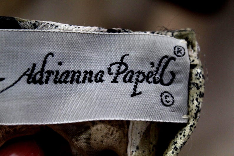 Pu&ograve; includere: Un'etichetta rettangolare per abbigliamento di colore grigio chiaro con il nome del marchio Adrianna Papell stampato in corsivo nero. L'etichetta &egrave; attaccata a un capo con un motivo testurizzato e maculato nei toni del nero, beige e bianco.