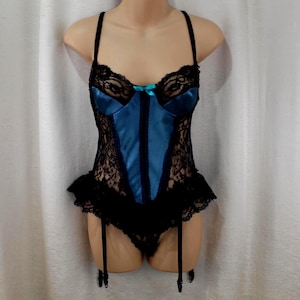 May include: A blue satin bodysuit featuring black lace overlays on the bodice and sides, a small turquoise bow at the center neckline, thin shoulder straps, a ruffled lace hemline, and attached garter straps hanging from the bottom edge.