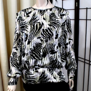 Pu&ograve; includere: Una camicetta a maniche lunghe con un motivo animalier nero e argento metallizzato simile a quello di una zebra su uno sfondo a righe verticali. Il capo presenta uno scollo rotondo, spalle leggermente scese e un orlo elasticizzato arricciato.