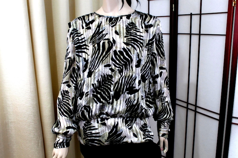 Pu&ograve; includere: Una camicetta a maniche lunghe con un motivo animalier nero e argento metallizzato simile a quello di una zebra su uno sfondo a righe verticali. Il capo presenta uno scollo rotondo, spalle leggermente scese e un orlo elasticizzato arricciato.