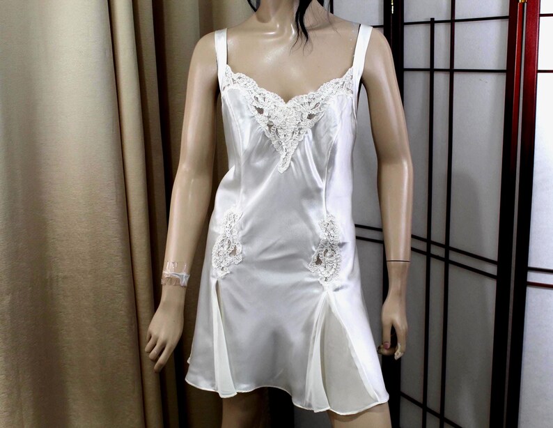 Puede incluir: Un camis&oacute;n de sat&eacute;n blanco con delicados adornos de encaje en el escote y el corpi&ntilde;o. El vestido presenta tirantes finos y detalles de encaje en la falda. El camis&oacute;n es una prenda de lencer&iacute;a cl&aacute;sica.