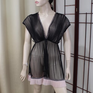 Op de afbeelding: Zwarte doorschijnende tuniek met een V-hals, kapmouwen en een geplooide zoom. De halslijn en zoom zijn afgezet met witte stippen. De tuniek wordt getoond op een mannequin.