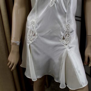 Puede incluir: Vestido lencero de sat&eacute;n blanco con detalles de encaje. El vestido presenta delicadas aplicaciones de encaje en el corpi&ntilde;o y la falda, con una abertura en el dobladillo. El vestido se exhibe en un maniqu&iacute;.