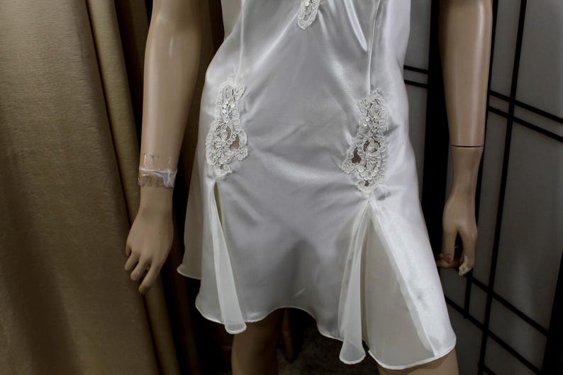 Puede incluir: Vestido lencero de sat&eacute;n blanco con detalles de encaje. El vestido presenta delicadas aplicaciones de encaje en el corpi&ntilde;o y la falda, con una abertura en el dobladillo. El vestido se exhibe en un maniqu&iacute;.