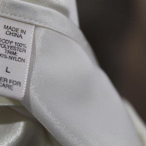 Puede incluir: Primer plano de una etiqueta de prenda blanca. La etiqueta dice "MADE IN CHINA" y detalla la composici&oacute;n de la tela: "BODY: 100% POLYESTER, TRIM: 100% NYLON". Tambi&eacute;n muestra la talla "L" y el texto "OVER FOR CARE".