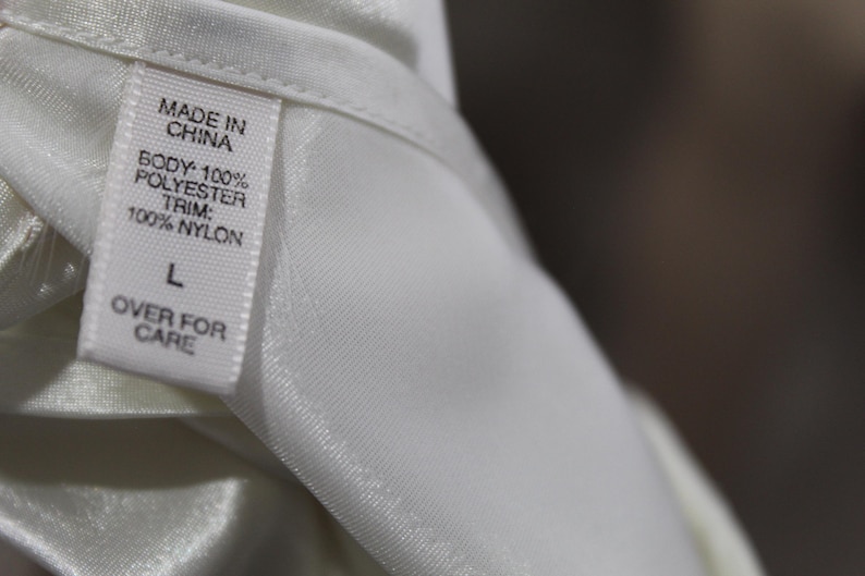 Puede incluir: Primer plano de una etiqueta de prenda blanca. La etiqueta dice "MADE IN CHINA" y detalla la composici&oacute;n de la tela: "BODY: 100% POLYESTER, TRIM: 100% NYLON". Tambi&eacute;n muestra la talla "L" y el texto "OVER FOR CARE".