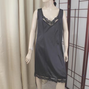 Op de afbeelding: Een zwarte slipdress met een V-hals en kanten afwerking. De mouwloze jurk is knielang en heeft delicate kanten details langs de halslijn en zoom. De jurk wordt getoond op een mannequin.