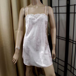 May include: A white satin slip dress with a floral pattern and lace trim. The dress has spaghetti straps and a knee-length hem. The mannequin is standing in front of a neutral background.