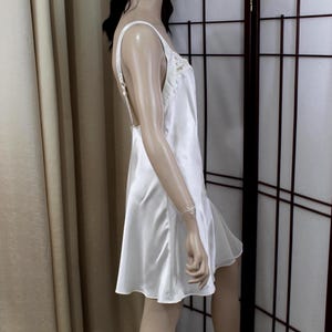 Puede incluir: Un vestido lencero de sat&eacute;n blanco con tirantes finos y espalda baja. El escote est&aacute; adornado con delicados detalles de encaje. El vestido tiene una falda acampanada y llega hasta la mitad del muslo. El vestido se muestra en un maniqu&iacute;.