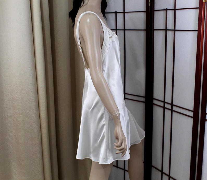 Puede incluir: Un vestido lencero de sat&eacute;n blanco con tirantes finos y espalda baja. El escote est&aacute; adornado con delicados detalles de encaje. El vestido tiene una falda acampanada y llega hasta la mitad del muslo. El vestido se muestra en un maniqu&iacute;.