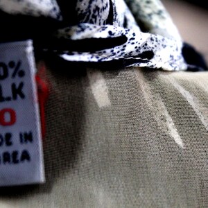 Pu&ograve; includere: Un'etichetta per abbigliamento in tessuto bianco con testo nero che riporta 100% SILK, il numero 10 in rosso e MADE IN KOREA. L'etichetta &egrave; attaccata a un capo con un motivo maculato bianco e nero e una sezione in tessuto beige chiaro trasparente.