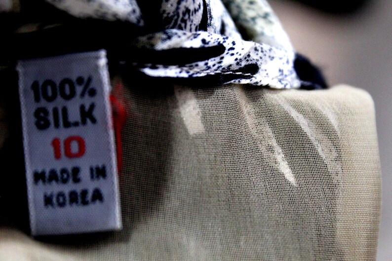 Pu&ograve; includere: Un'etichetta per abbigliamento in tessuto bianco con testo nero che riporta 100% SILK, il numero 10 in rosso e MADE IN KOREA. L'etichetta &egrave; attaccata a un capo con un motivo maculato bianco e nero e una sezione in tessuto beige chiaro trasparente.