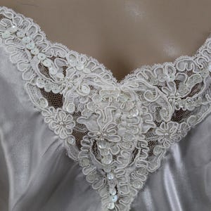 Puede incluir: Primer plano de una camisola de sat&eacute;n blanco con delicados adornos de encaje. El escote en V est&aacute; adornado con intrincado encaje floral, adornado con lentejuelas y cuentas. La tela suave cae suavemente, creando un aspecto rom&aacute;ntico y elegante.