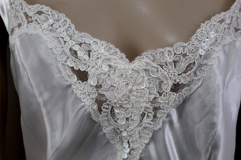 Puede incluir: Primer plano de una camisola de sat&eacute;n blanco con delicados adornos de encaje. El escote en V est&aacute; adornado con intrincado encaje floral, adornado con lentejuelas y cuentas. La tela suave cae suavemente, creando un aspecto rom&aacute;ntico y elegante.