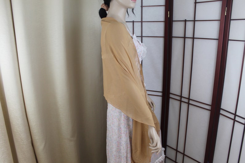 May include: A light brown sheer scarf draped over a person's shoulders. The scarf is made of a soft, flowing fabric and has a simple, elegant design.