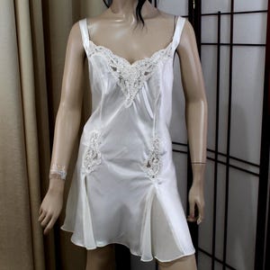 Puede incluir: Un camis&oacute;n de sat&eacute;n blanco con delicados adornos de encaje. El vestido presenta tirantes finos, un escote en V y detalles de encaje en el corpi&ntilde;o y la falda. El vestido tiene una abertura a cada lado, a&ntilde;adiendo un toque de elegancia.