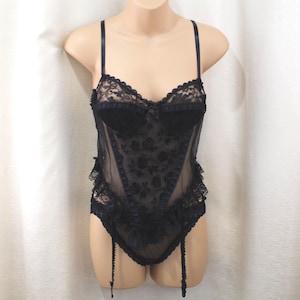 May include: A black lace bodysuit featuring a floral pattern, thin adjustable shoulder straps, and a sweetheart neckline. The garment includes structured cups, sheer mesh panels, ruffled lace trim along the sides, and attached garter straps.