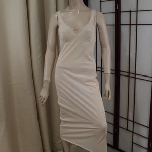 Op de afbeelding: Een beige onderjurk met een V-hals en delicate kanten afwerking. De mouwloze jurk valt tot halverwege de kuit. De mannequin draagt een bijpassende kanten armband. De jurk is gemaakt van een zachte, vloeiende stof.