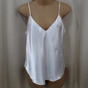 Può includere: Una camisole di raso bianco con scollo a V e spalline sottili. La parte superiore ha una vestibilità ampia e un orlo curvo. Questo capo di lingerie è esposto su un manichino.