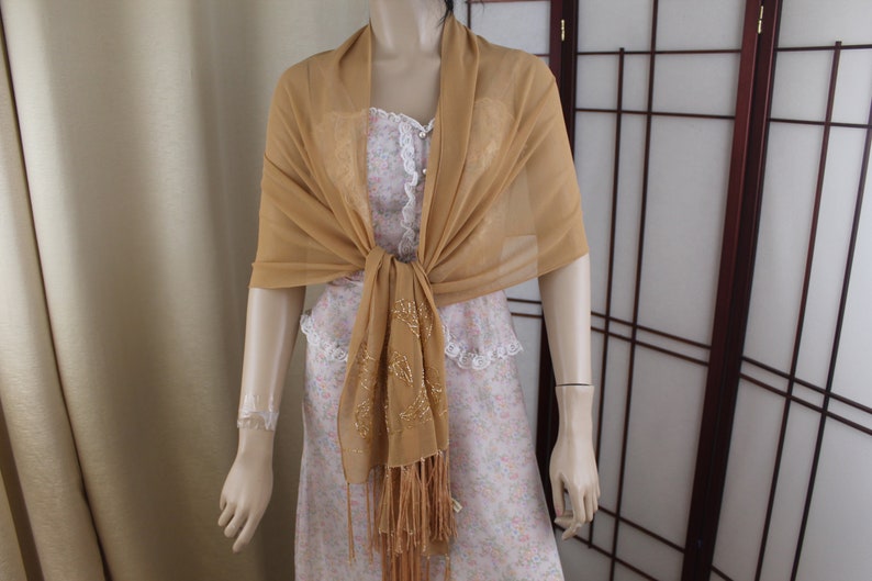 May include: A gold sheer shawl with a beaded fringe. The shawl is draped over a mannequin and tied at the neck.