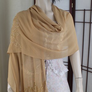 May include: A beige chiffon shawl with gold leaf embroidery. The shawl is draped over a mannequin's shoulders.