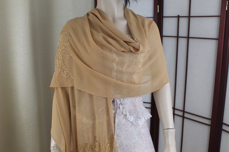 May include: A beige chiffon shawl with gold leaf embroidery. The shawl is draped over a mannequin's shoulders.