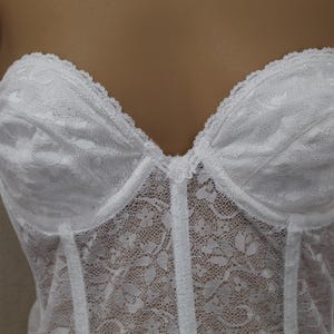 May include: A white strapless corset top featuring an intricate floral lace overlay. The garment has structured, molded cups with a scalloped lace trim along the neckline and a small decorative floral detail at the center of the bust.