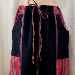 May include: Black trousers featuring a high elastic waistband with a drawstring tie. The pants are designed with vertical panels of multi-colored striped fabric in shades of red, pink, and blue, contrasting with solid black sections and side pockets.