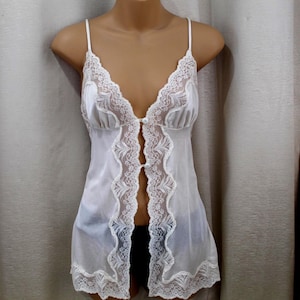 Könnte beinhalten: Weißes Dessous-Top mit zarten Spitzenbesatz und Spaghettiträgern. Die offene Vorderseite hat dekorative Spitzeneinsätze und kleine Knöpfe. Das Kleidungsstück wird auf einer Schaufensterpuppe präsentiert.
