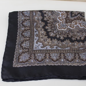 May include: A folded black silk scarf with an intricate paisley pattern in shades of brown, white, and blue. The scarf has a solid black border and a detailed design, suitable for adding a touch of elegance to any outfit.