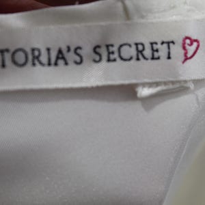 Puede incluir: Primer plano de una etiqueta de tela blanca con el texto "VICTORIA'S SECRET" en negro y un s&iacute;mbolo de coraz&oacute;n rosa. La etiqueta est&aacute; cosida sobre una tela de sat&eacute;n blanca.